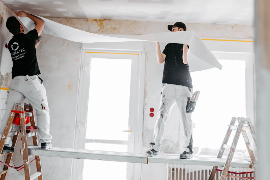 Renovierungsarbeiten eines Raumes: Zwei Handwerker mit schwarzen T-Shirts und weißen Hosen tapezieren eine Decke.